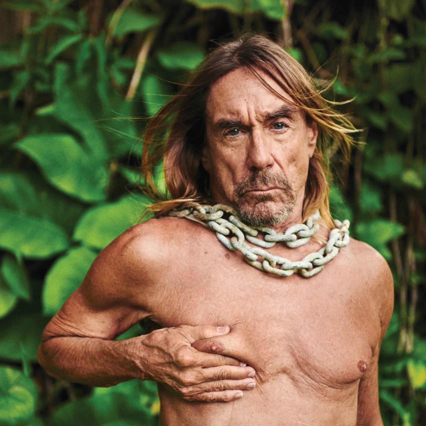 Iggy Pop standing in lush green foliage wearing a heavy chain necklace